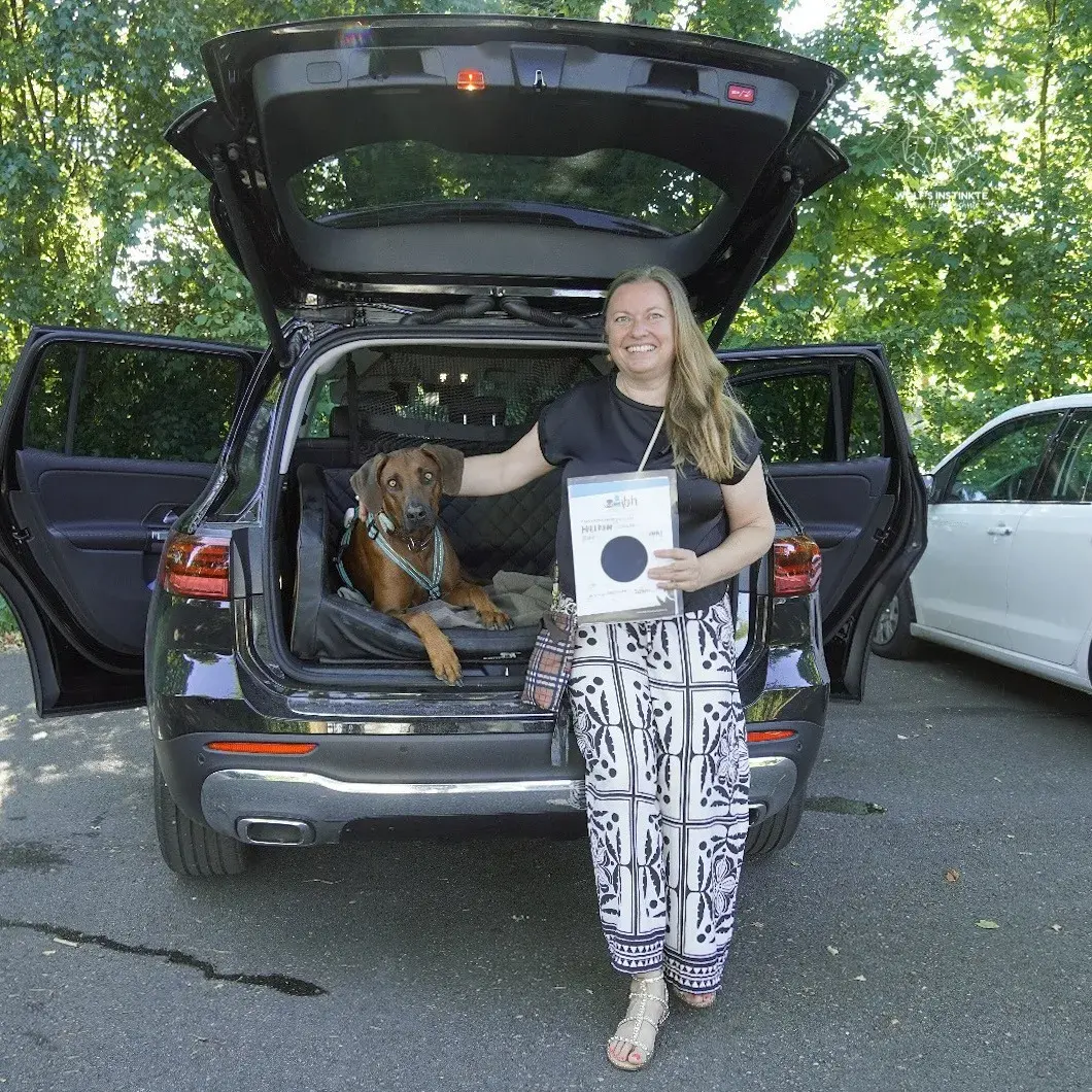 Teilnehmerin mit Ridgeback und Prüfungsurkunde am geöffneten Kofferraum
